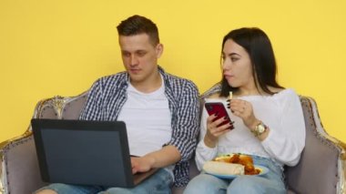 Adam ve kız fast food yiyor, adam dizüstü bilgisayarın başında oturuyor ve kız da sarı arka plandaki telefona bakıyor. Sağlıklı ve sağlıksız gıda kavramı. Fast food