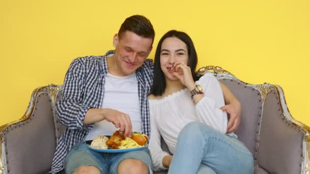 Un ragazzo e una ragazza mangiano fast food seduti su un divano su uno sfondo giallo. coppia guardando la fotocamera. Il concetto di cibo sano e malsano. Fast food — Video Stock