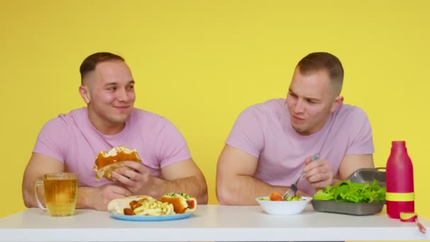 दोन स्नायू जुळे पिवळ्या पार्श्वभूमीवर टेबलावर निरोगी अन्न आणि वेगवान अन्न खातात. निरोगी आणि निरोगी अन्नाची संकल्पना. फास्ट फूड — स्टॉक व्हिडिओ