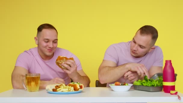 Δύο μυώδεις δίδυμοι τρώνε υγιεινό φαγητό και γρήγορο φαγητό στο τραπέζι σε κίτρινο φόντο. Η έννοια των υγιεινών και ανθυγιεινών τροφίμων. Γρήγορο φαγητό — Αρχείο Βίντεο