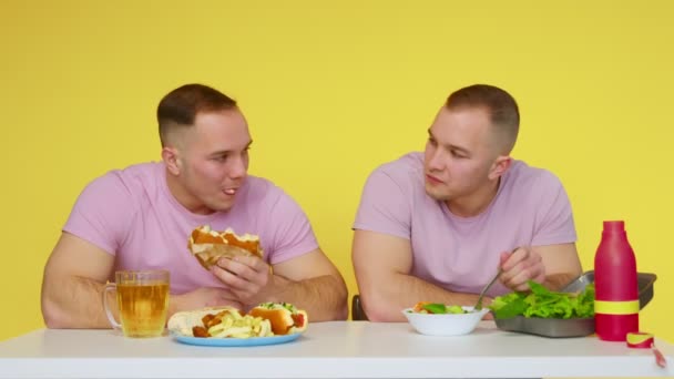 Dos gemelos musculosos comen comida saludable y comida rápida en la mesa sobre un fondo amarillo. El concepto de comida saludable y poco saludable. Comida rápida — Vídeos de Stock