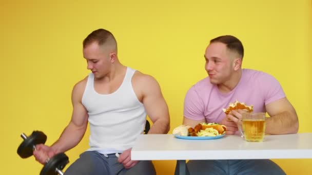 Два м'язистих близнюка на жовтому тлі. Один хлопець гойдається з гантелями, а другий їсть фаст-фуди. Концепція здорової та здорової їжі. фаст-фуд — стокове відео