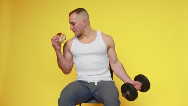 Un ragazzo muscoloso solleva un manubrio con una mano e un hamburger con l'altra mano. Il concetto di cibo sano e malsano. — Video Stock