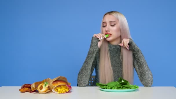 Une jolie fille mange des légumes verts et regarde tristement la restauration rapide sur un fond bleu. Régime alimentaire. Le concept d'aliments sains et malsains. Restauration rapide — Video