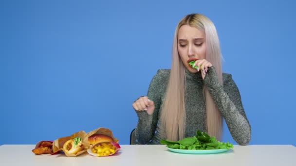 Una ragazza carina mangia verdure e guarda tristemente fast food su uno sfondo blu. Dieta. Il concetto di cibo sano e malsano. Fast food — Video Stock