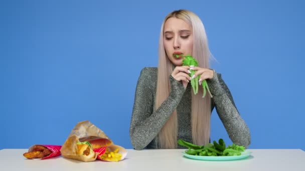 Симпатичная девушка ест зелень и грустно смотрит на фаст-фуд на синем фоне. Девушка прерывает диету и ест фаст-фуд. Диета. Понятие здоровой и нездоровой пищи. Фаст-фуд — стоковое видео