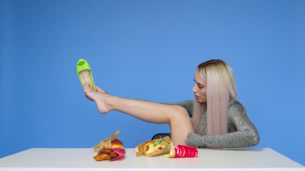 Słodka dziewczyna siedzi na krześle, zdejmuje buty, kładzie nogi na stole i zaczyna jeść hamburgera na niebieskim tle.Diet. Koncepcja zdrowej i niezdrowej żywności. Fast food — Wideo stockowe
