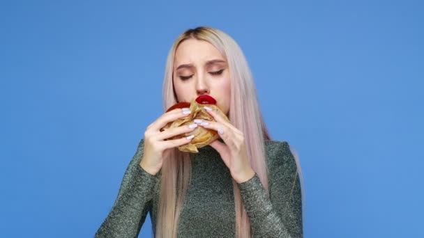 Närbild av en flicka med rött läppstift på läpparna, äter en hamburgare och smetar läppstift med handen på en blå bakgrund. Föda. Begreppet hälsosam och ohälsosam mat. snabbmat — Stockvideo