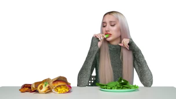 Uma garota bonita come verduras e olha tristemente para fast food em um fundo branco. Dieta. O conceito de comida saudável e insalubre. fast food — Vídeo de Stock