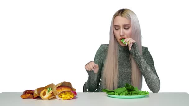 Una linda chica come verduras y mira tristemente la comida rápida sobre un fondo blanco. Dieta. El concepto de comida saludable y poco saludable. comida rápida — Vídeo de stock