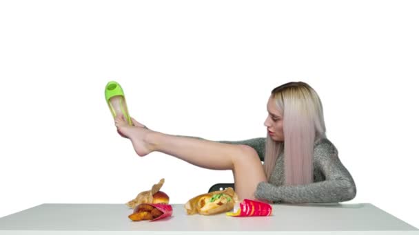 Une jolie fille s'assoit sur une chaise, enlève ses chaussures, pose ses pieds sur la table et commence à manger un hamburger sur un fond blanc. Régime alimentaire. Le concept d'aliments sains et malsains. Restauration rapide — Video