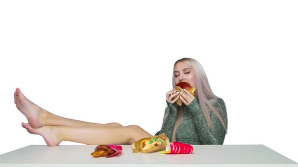 En söt flicka sitter på en stol med benen på bordet och äter en hamburgare på en vit bakgrund. Föda. Begreppet hälsosam och ohälsosam mat. snabbmat — Stockvideo
