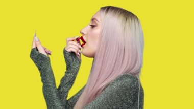 Bir kız dudaklarını kırmızı rujla boyuyor ve sarı arka planda zevkle hamburger yemeye başlıyor. Diyet olsun. Sağlıklı ve sağlıksız gıda kavramı. fast food