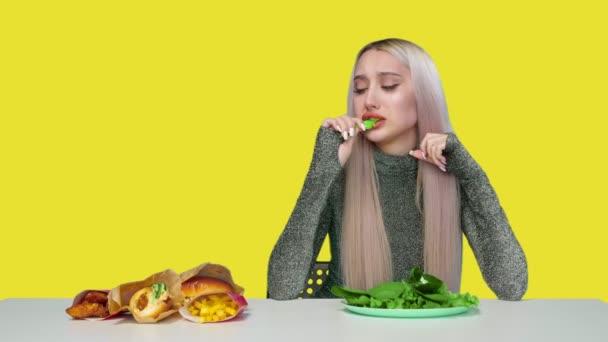 かわいい女の子は緑を食べ、悲しいことに黄色の背景にファーストフードを見ています。食事よ。健康的で不健康な食べ物の概念。ファーストフード — ストック動画