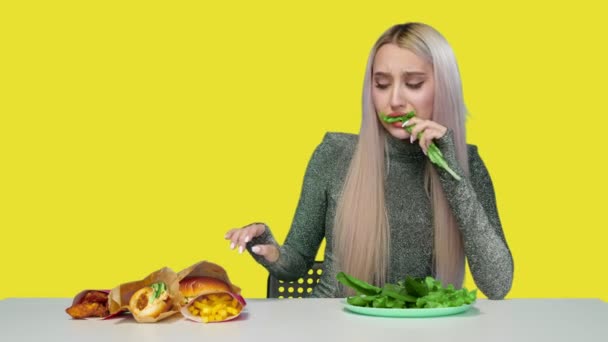 Uma garota bonita come verduras e olha tristemente para fast food em um fundo amarelo. Dieta. O conceito de comida saudável e insalubre. fast food — Vídeo de Stock
