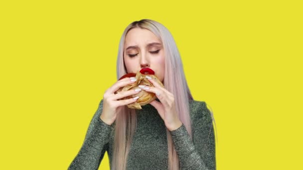 Gros plan d'une fille avec du rouge à lèvres rouge sur les lèvres, mangeant un hamburger et enduisant de rouge à lèvres avec sa main sur un fond jaune. Régime alimentaire. Le concept d'aliments sains et malsains. restauration rapide — Video