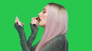 Bir kız dudaklarını kırmızı rujla boyuyor ve yeşil arka planda zevkle hamburger yemeye başlıyor. Diyet olsun. Sağlıklı ve sağlıksız gıda kavramı. fast food