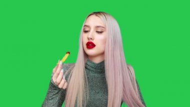 Bir kız dudaklarını kırmızı rujla boyuyor ve yeşil arka planda zevkle hamburger yemeye başlıyor. Diyet olsun. Sağlıklı ve sağlıksız gıda kavramı. fast food