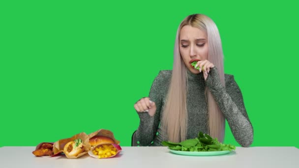 かわいい女の子は緑を食べ、悲しいことに緑の背景にファーストフードを見ています。食事よ。健康的で不健康な食べ物の概念。ファーストフード — ストック動画