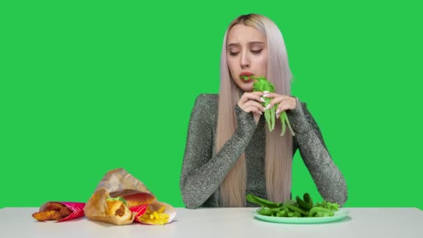 Une jolie fille mange des légumes verts et regarde tristement la restauration rapide sur un fond vert. La fille interrompt le régime alimentaire et mange de la restauration rapide. Régime alimentaire. Le concept d'aliments sains et malsains. restauration rapide — Video