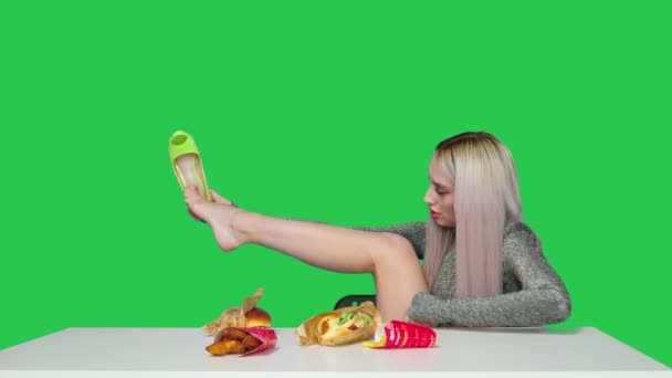 한 귀여운 소녀가 의자에 앉아 신발을 벗고 탁자 위에 발을 올려 녹색 배경에 있는 햄버거를 먹기 시작 한다. 다이어트. 건강하고 건강하지 못한 음식의 개념. 패스트푸드 — 비디오