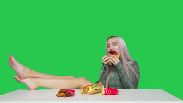 O fată drăguță stă pe un scaun cu picioarele pe masă și mănâncă un hamburger pe un fundal verde. Dieta. Conceptul de alimente sănătoase și nesănătoase. fast food — Videoclip de stoc