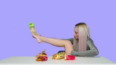 Şirin bir kız sandalyeye oturur, ayakkabılarını çıkarır, ayaklarını masaya koyar ve mor bir arka planda hamburger yemeye başlar. Diyet olsun. Sağlıklı ve sağlıksız gıda kavramı. fast food