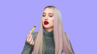 Bir kız dudaklarını kırmızı rujla boyuyor ve mor arka planda zevkle hamburger yemeye başlıyor. Diyet olsun. Sağlıklı ve sağlıksız gıda kavramı. fast food