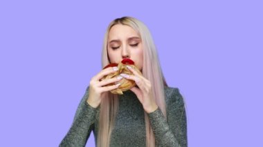 Dudaklarında kırmızı ruj lekesi olan, hamburger yiyen ve elini mor arka plana sürerek ruj süren bir kızın yakın çekimi. Diyet olsun. Sağlıklı ve sağlıksız gıda kavramı. fast food