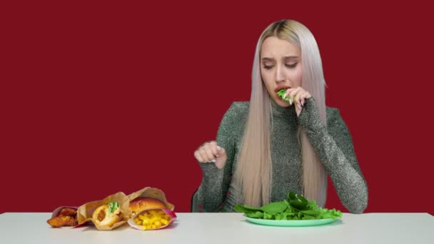 Симпатичная девушка ест зелень и печально смотрит на фаст-фуд на красном фоне. Диета. Понятие здоровой и нездоровой пищи. фастфуд — стоковое видео