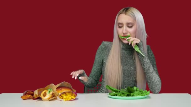 Egy aranyos lány zöldséget eszik, és szomorúan nézi a gyorskaját vörös alapon. Fogyókúra. Az egészséges és egészségtelen ételek fogalma. gyorsétterem — Stock videók