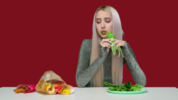 Tatlı bir kız yeşillik yer ve kırmızı arka plandaki fast food 'a üzülerek bakar. Kız diyeti bozuyor ve fast food yiyor. Diyet olsun. Sağlıklı ve sağlıksız gıda kavramı. fast food — Stok video