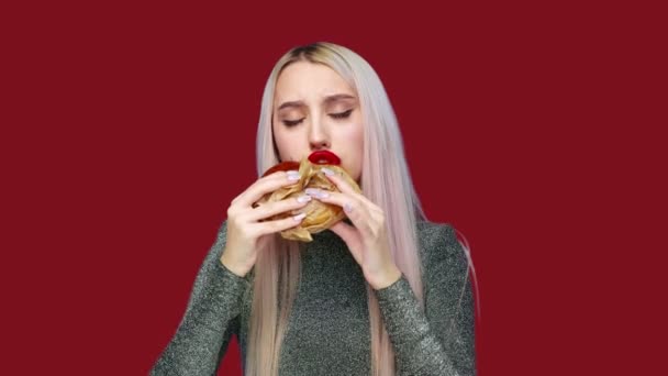Närbild av en flicka med rött läppstift på läpparna, äta en hamburgare och smeta läppstift med handen på en röd bakgrund. Föda. Begreppet hälsosam och ohälsosam mat. snabbmat — Stockvideo