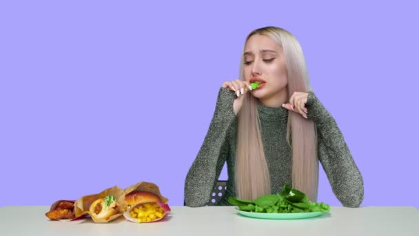 Una linda chica come verduras y mira tristemente la comida rápida sobre un fondo púrpura. Dieta. El concepto de comida saludable y poco saludable. comida rápida — Vídeo de stock