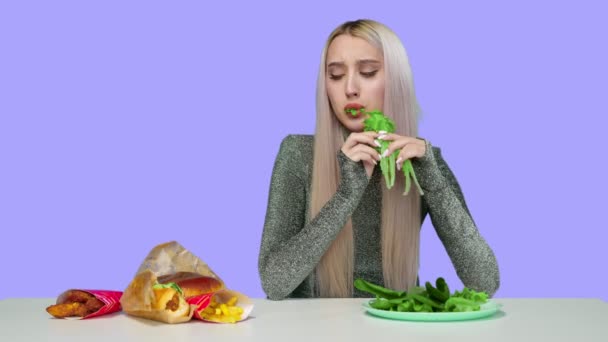 Roztomilá dívka jí zeleninu a smutně se dívá na fast food na fialovém pozadí. Dívka přerušuje dietu a jí rychlé občerstvení. Dieta. Koncept zdravé a nezdravé stravy. fast food — Stock video