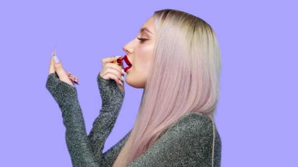 Närbild av en flicka målar sina läppar med rött läppstift och börjar äta en hamburgare med glädje på en lila bakgrund. Föda. Begreppet hälsosam och ohälsosam mat. snabbmat — Stockvideo