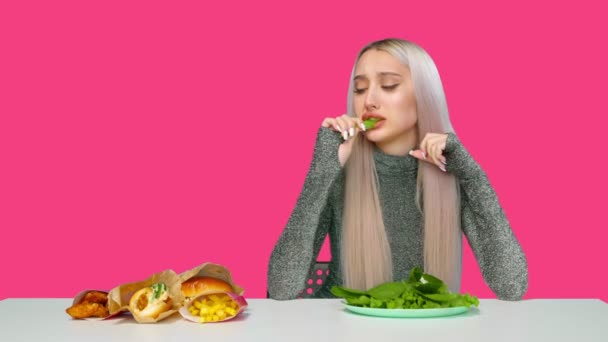 一个可爱的女孩吃着蔬菜，忧郁地看着粉色背景下的快餐。节食。健康和不健康食品的概念。快餐食品 — 图库视频影像
