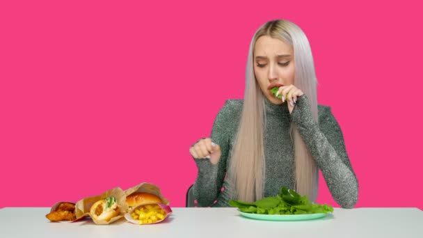 Roztomilé děvče jí zeleninu a smutně se dívá na fast food na růžovém pozadí. Dieta. Koncept zdravé a nezdravé stravy. fast food — Stock video