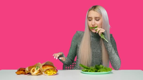 Egy aranyos lány zöldséget eszik, és szomorúan nézi a gyorskaját rózsaszín alapon. Fogyókúra. Az egészséges és egészségtelen ételek fogalma. gyorsétterem — Stock videók