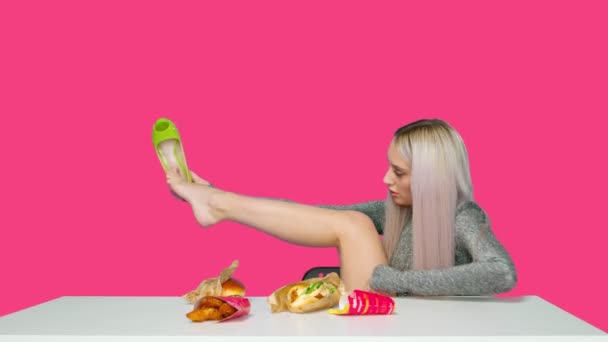 Słodka dziewczyna siedzi na krześle, zdejmuje buty, kładzie nogi na stole i zaczyna jeść hamburgera na różowym tle. Dieta. Koncepcja zdrowej i niezdrowej żywności. fast food — Wideo stockowe
