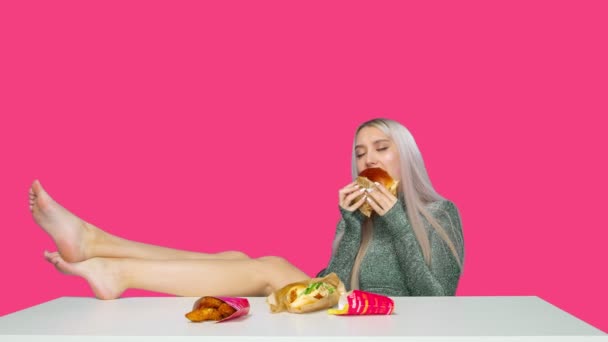 Симпатичная девушка сидит на стуле с ногами на столе и ест гамбургер на розовом фоне. Диета. Понятие здоровой и нездоровой пищи. фастфуд — стоковое видео