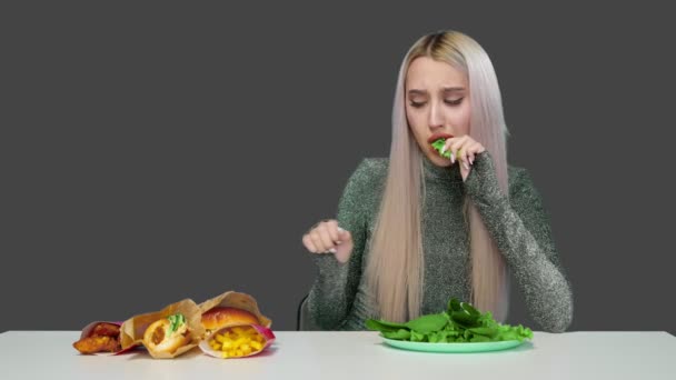 Une jolie fille mange des légumes verts et regarde tristement la restauration rapide sur un fond gris. Régime alimentaire. Le concept d'aliments sains et malsains. restauration rapide — Video