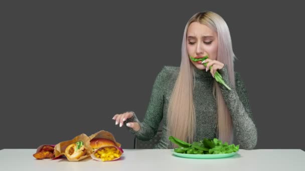 Una ragazza carina mangia verdure e purtroppo guarda fast food su uno sfondo grigio. Dieta. Il concetto di cibo sano e malsano. fast food — Video Stock