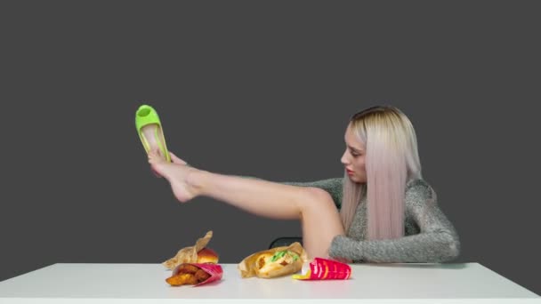 Ein nettes Mädchen sitzt auf einem Stuhl, zieht die Schuhe aus, stellt die Füße auf den Tisch und beginnt einen Hamburger auf grauem Hintergrund zu essen. Ernährung. Das Konzept der gesunden und ungesunden Ernährung. fast food — Stockvideo