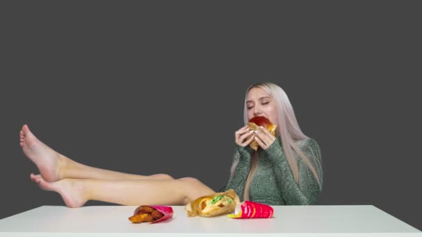 Una ragazza carina siede su una sedia con le gambe sul tavolo e mangia un hamburger su uno sfondo grigio. Dieta. Il concetto di cibo sano e malsano. fast food — Video Stock