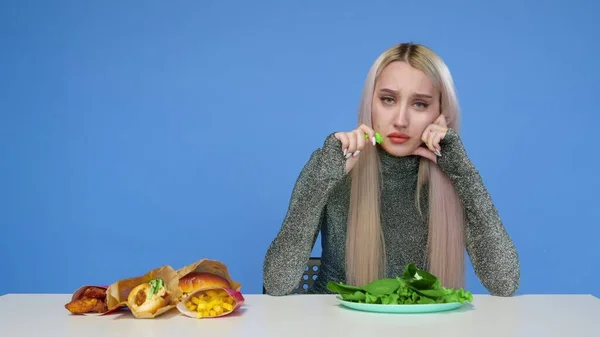 Мила дівчина їсть зелень і, на жаль, виглядає на фаст-фуді на синьому фоні. Дієта. Концепція здорової та здорової їжі. Фастфуд — стокове фото