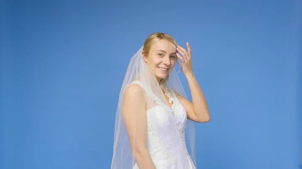 Young beautiful blonde smiling in a wedding white dress and veil on a blue background. The girl looks into the camera. Wedding — Stock Photo, Image