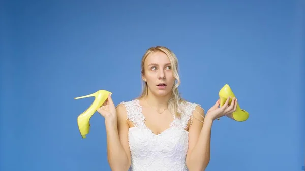 Gefolterte junge schöne blonde Frau in einem weißen Hochzeitskleid und Schleier auf blauem Grund, zieht ihre Schuhe aus. Das Mädchen blickt in die Kamera. Hochzeit — Stockfoto