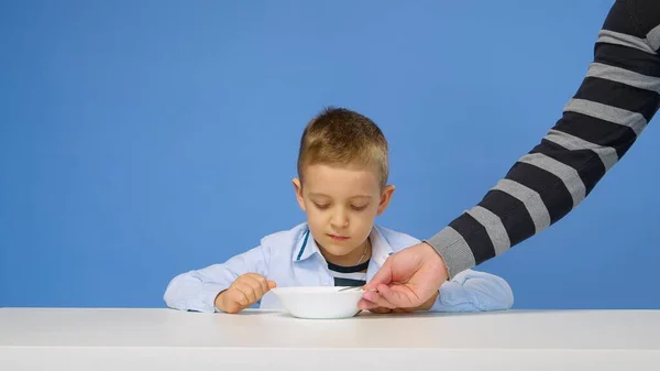 Ein Knabenkind sitzt am Tisch und will vor blauem Hintergrund keinen Haferbrei essen. Trauriges und unzufriedenes Gesicht. Konzept für gesundes Frühstück — Stockfoto
