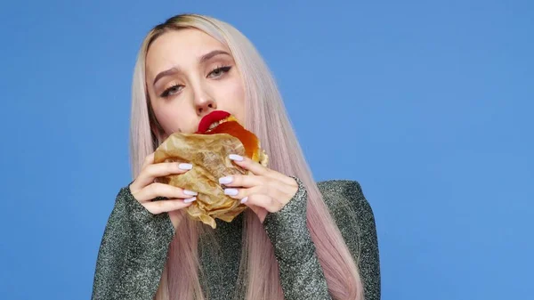 Close-up de uma menina com batom vermelho nos lábios, comendo um hambúrguer em um fundo azul. Dieta. O conceito de comida saudável e insalubre. fast food — Fotografia de Stock